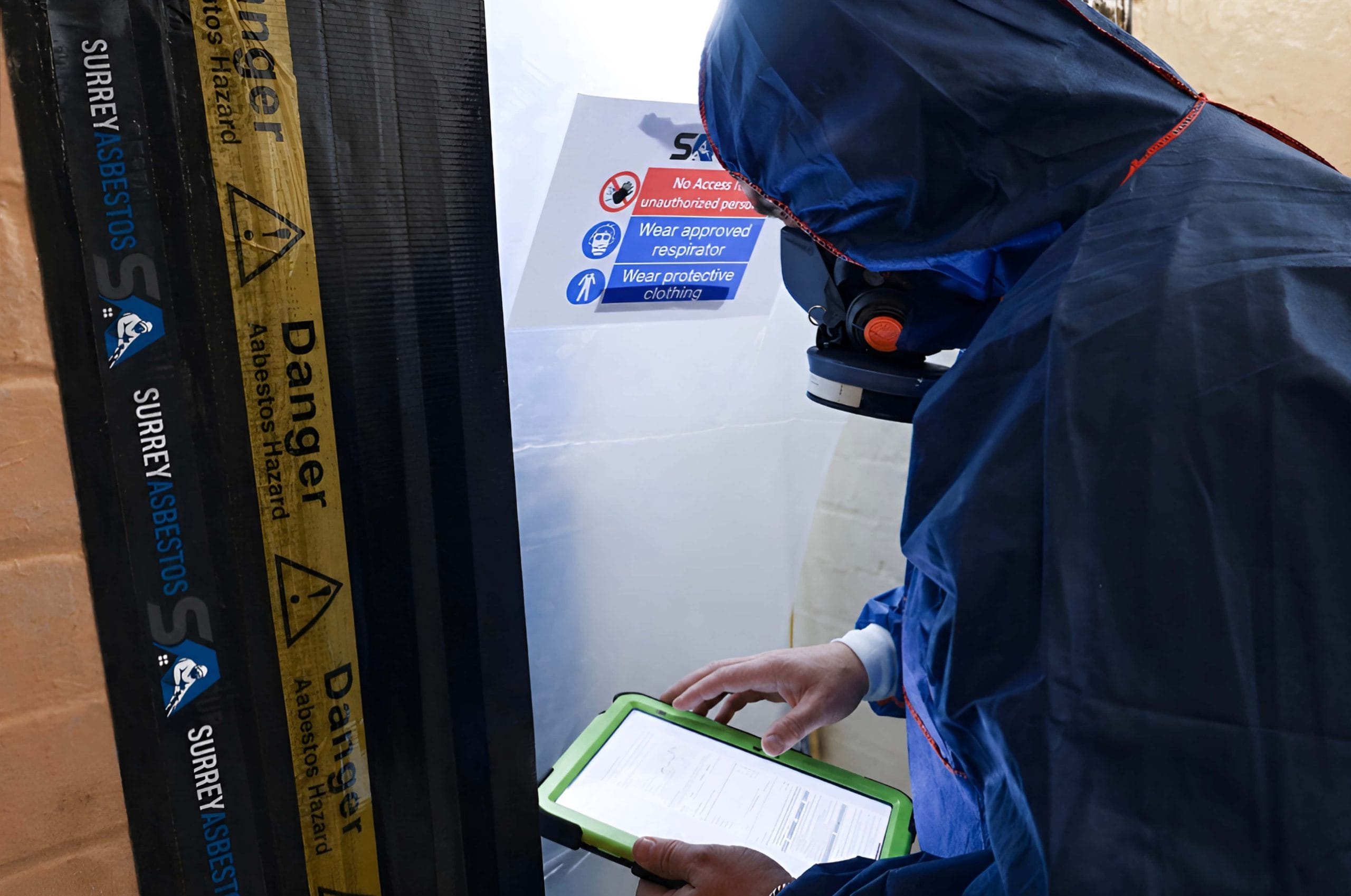 Surrey Asbestos operative in full PPE reviewing documentation at a sealed asbestos hazard enclosure entry point in Surrey