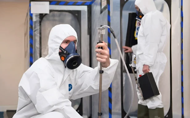 An asbestos analyst in full protective coveralls and respiratory mask setting up air monitoring equipment next to a sealed enclosure on a Surrey job site.